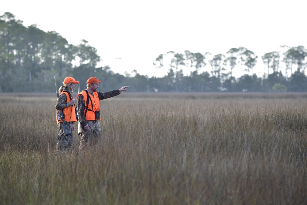 Two hunters wearing blaze-orange vests and caps stand in tall grass, with one pointing across the field.