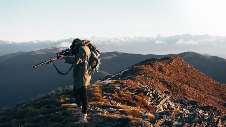 Hunter carrying a backpack and rifle on a high mountain ridge at sunrise.