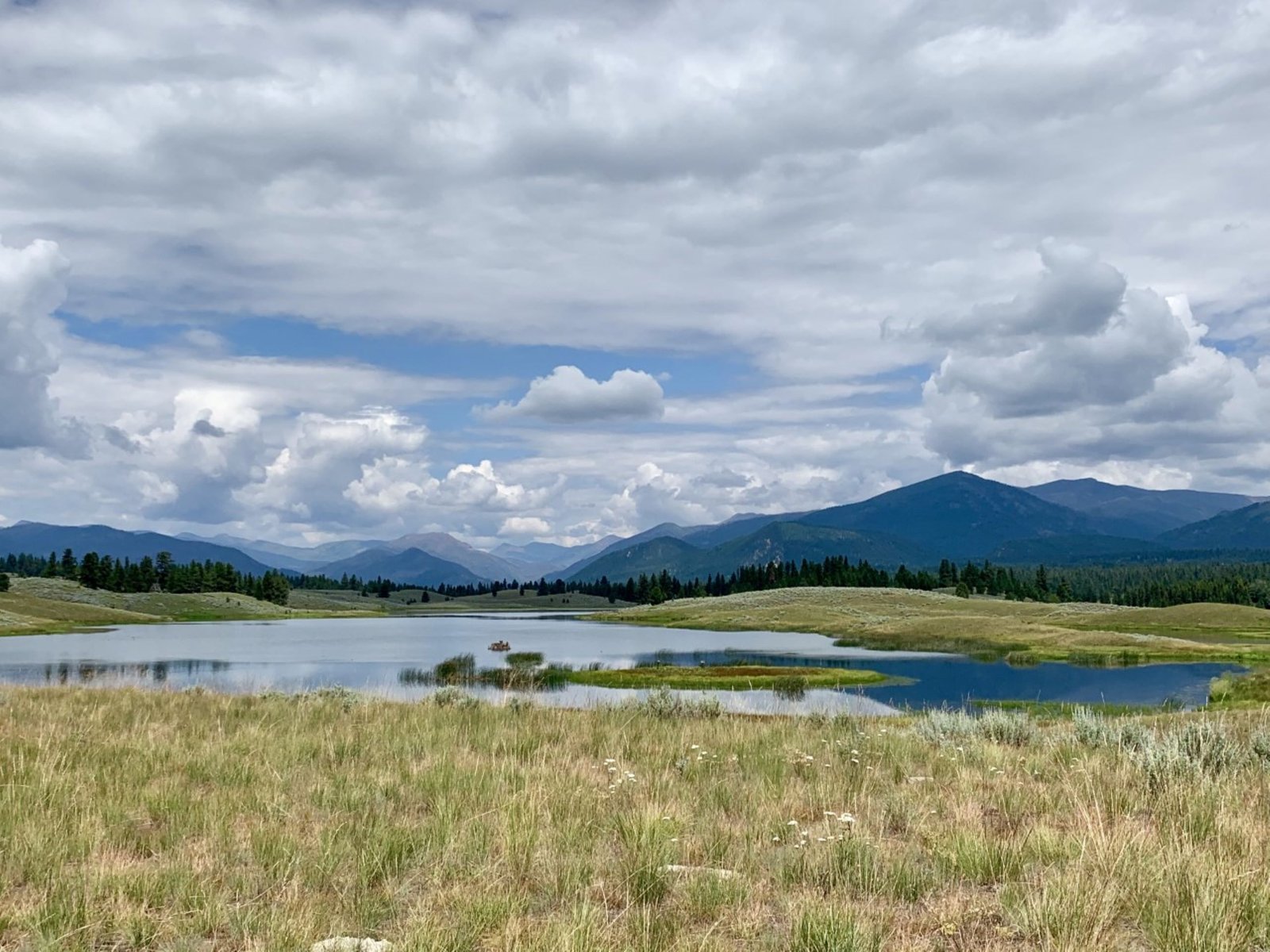 A Montana lake bordered by open grassland and forested mountains beneath layered clouds